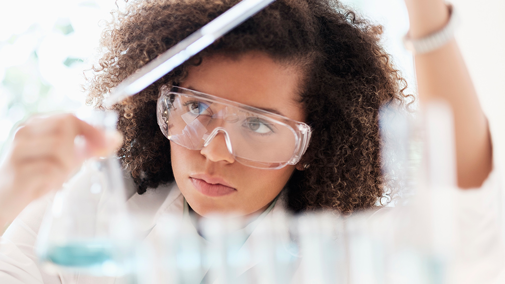 A female scientist with goggles in her lab examining substances in beakers.