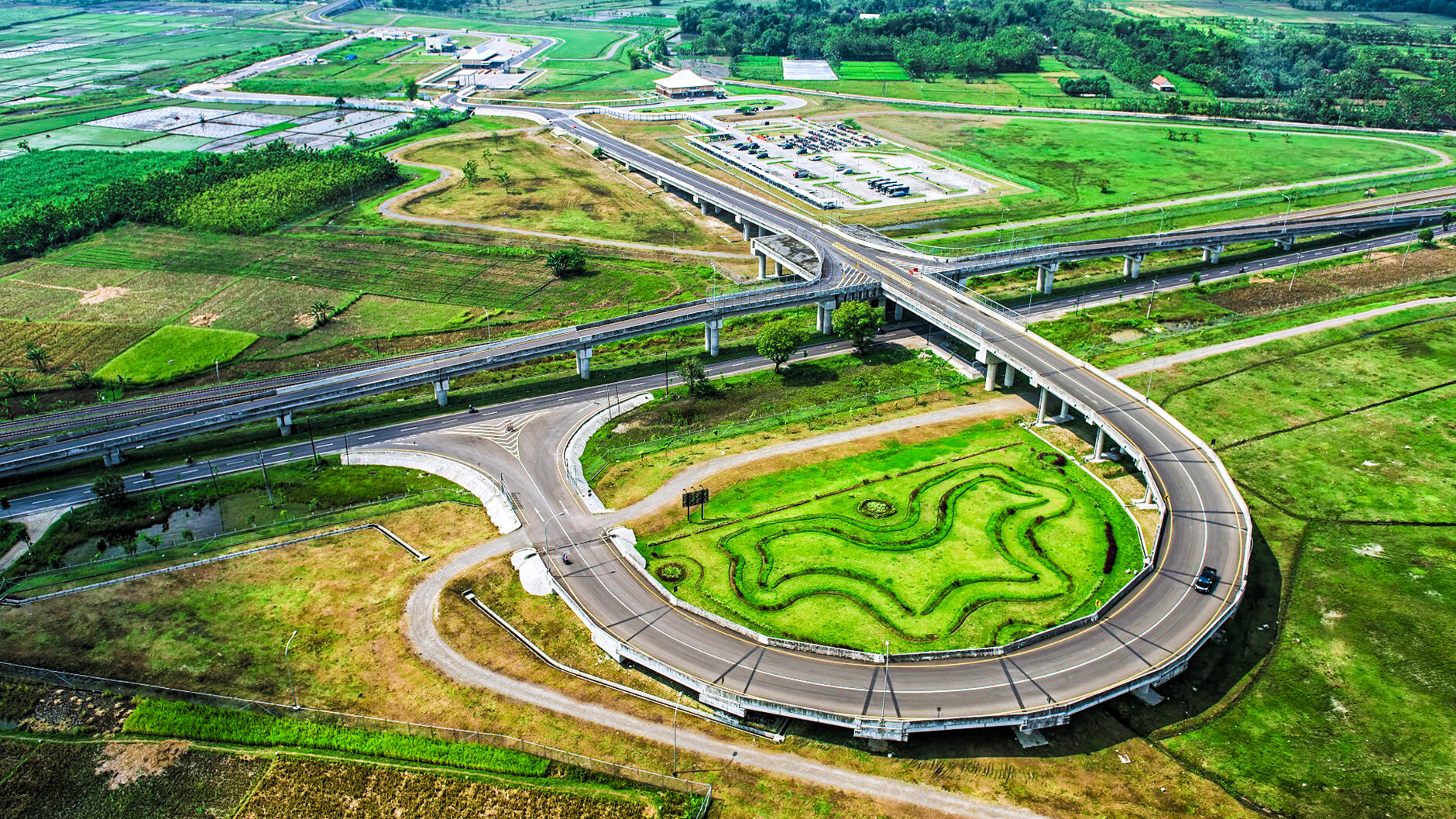 Aerial view of a curved highway interchange surrounded by green fields and farmlands, with a decorative green landscaped area in the center of the loop.