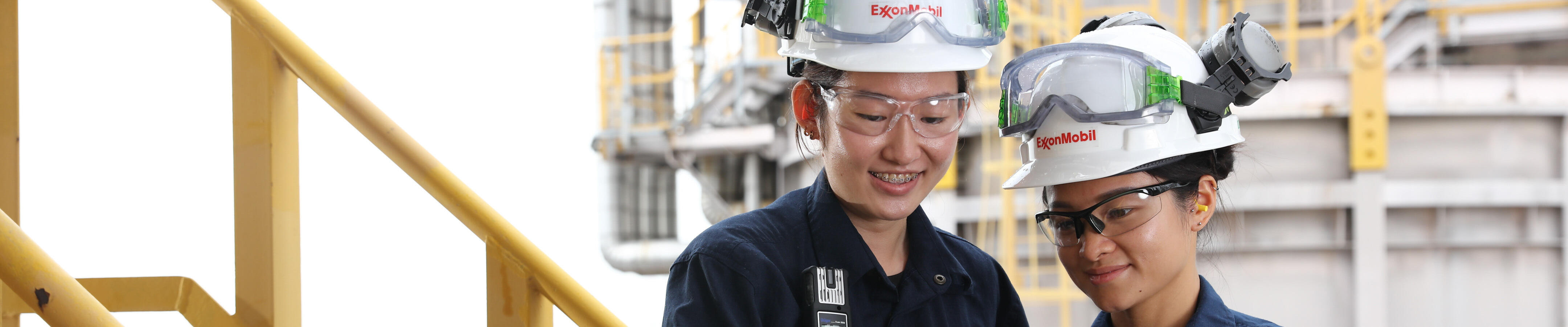 Two workers wearing navy blue coveralls and white safety helmets with the ExxonMobil logo, standing on a yellow metal staircase in an industrial setting. Both workers are equipped with safety goggles and appear to be engaged in a discussion while looking at a tablet device. The background includes industrial equipment and structures, with greenery visible beyond the facility. The environment suggests they are on a site related to energy, chemicals, or manufacturing, and they are likely reviewing operational or safety procedures.
