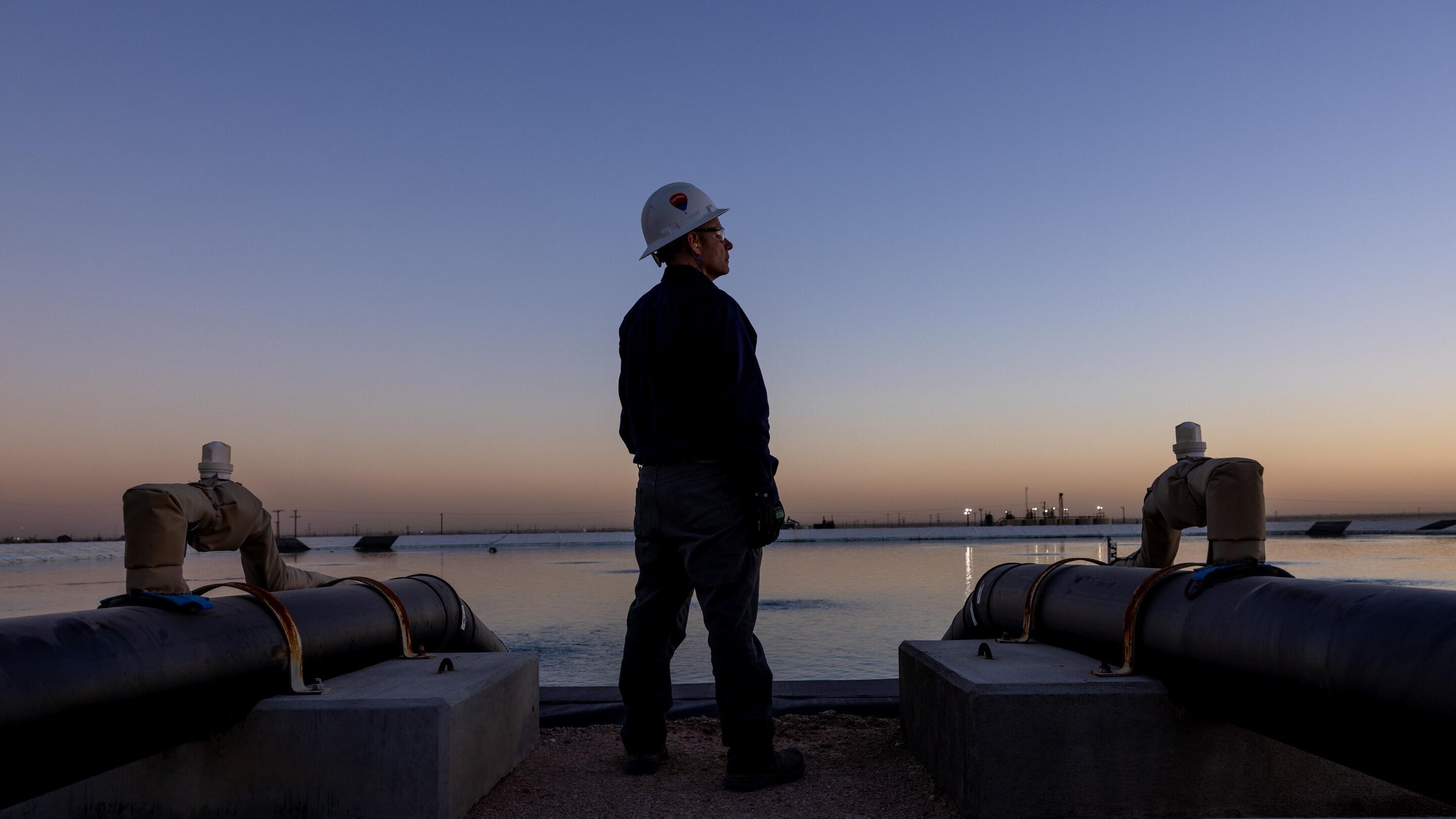The image depicts a worker standing between two large industrial pipes near a body of water during twilight. The worker is wearing a hard hat and gloves, suggesting they are engaged in a task related to construction or maintenance. The pipes are anchored on concrete bases and extend toward the water. In the background, the sky displays a gradient of colors as the sun sets, and distant structures can be seen along the horizon. The scene conveys an atmosphere of industrial activity set against a serene natural backdrop.