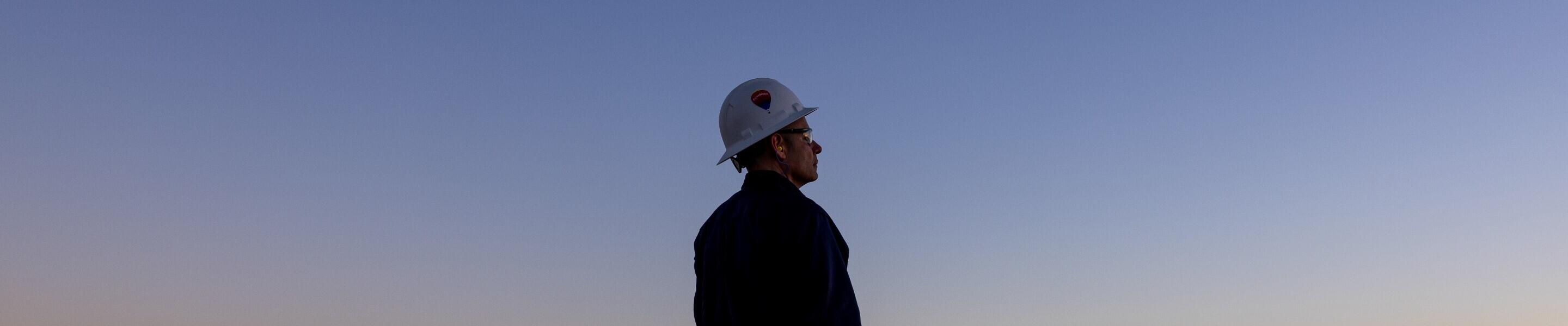 The image depicts a worker standing between two large industrial pipes near a body of water during twilight. The worker is wearing a hard hat and gloves, suggesting they are engaged in a task related to construction or maintenance. The pipes are anchored on concrete bases and extend toward the water. In the background, the sky displays a gradient of colors as the sun sets, and distant structures can be seen along the horizon. The scene conveys an atmosphere of industrial activity set against a serene natural backdrop.