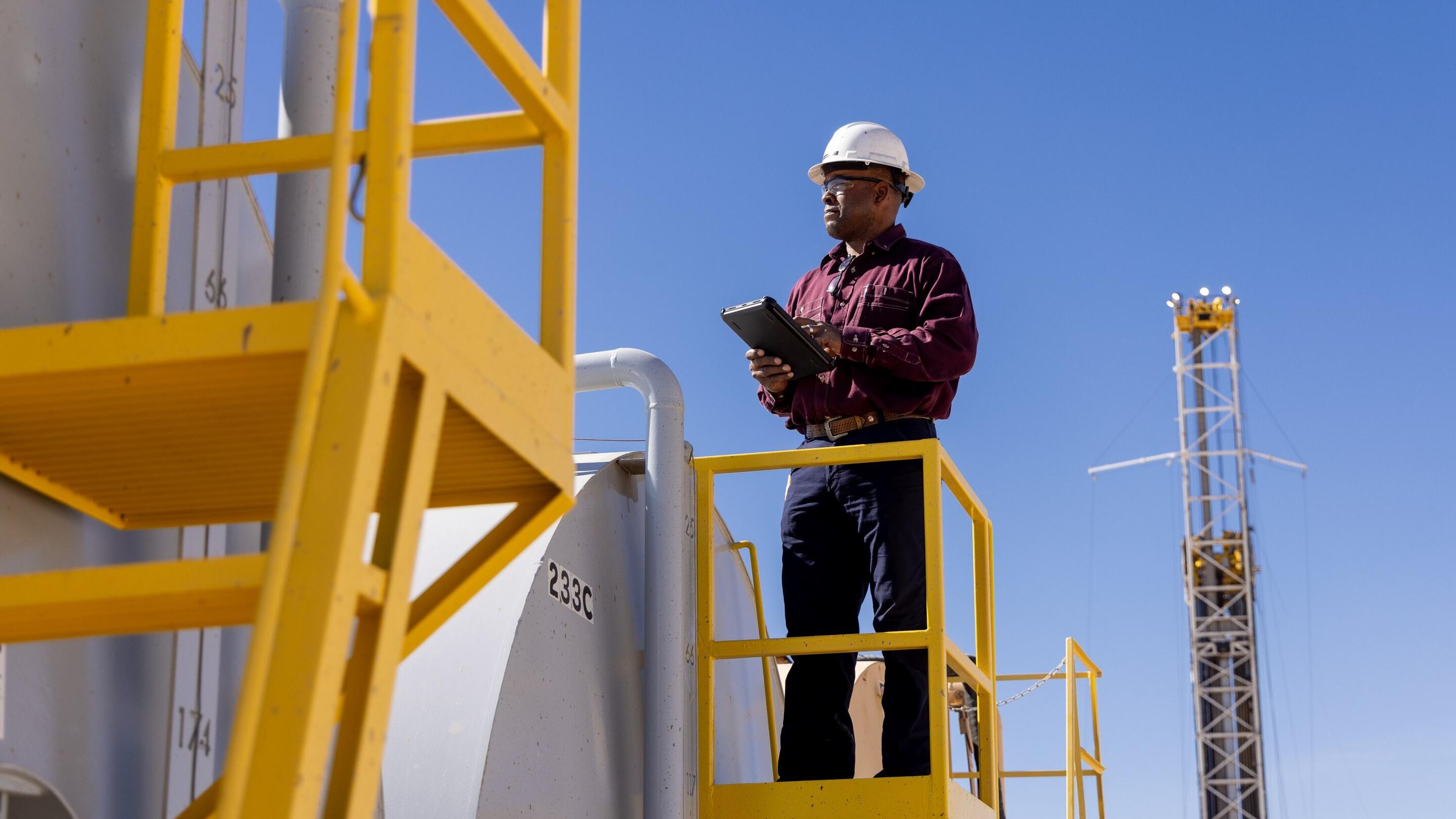A person wearing a white hard hat and a maroon long-sleeve shirt standing on a yellow metal platform. They are holding a tablet, possibly conducting an inspection or taking notes. Behind them, there are large industrial tanks, and a tall structure, possibly a drilling rig, is visible in the background against a clear blue sky. The scene appears to be set in an industrial facility, likely related to the oil or gas industry. The use of safety equipment and structured walkways suggests adherence to safety protocols.
