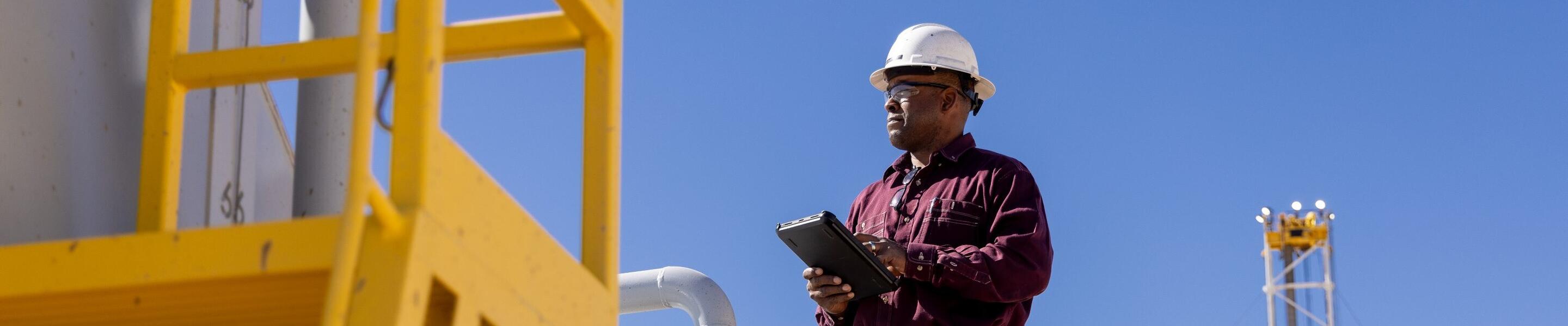 A person wearing a white hard hat and a maroon long-sleeve shirt standing on a yellow metal platform. They are holding a tablet, possibly conducting an inspection or taking notes. Behind them, there are large industrial tanks, and a tall structure, possibly a drilling rig, is visible in the background against a clear blue sky. The scene appears to be set in an industrial facility, likely related to the oil or gas industry. The use of safety equipment and structured walkways suggests adherence to safety protocols.