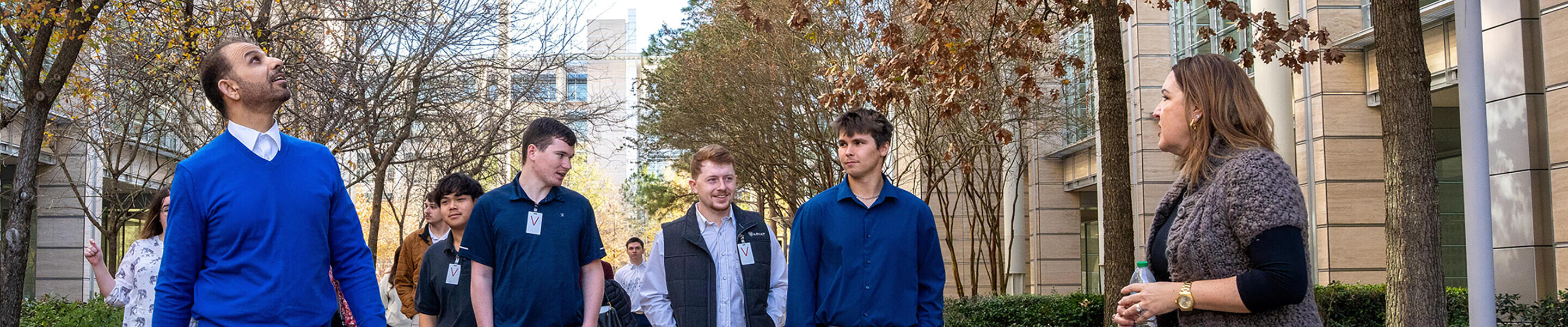 A group of people walking outside in what appears to be a campus or office park setting. Several individuals are wearing business casual attire, including sweaters, collared shirts, and vests, and some have visible name tags or badges. The setting is lined with trees, some of which have autumn-colored leaves, and there are modern buildings in the background. One person on the right is holding a water bottle and seems to be speaking to the group, possibly leading a tour or discussion. The overall atmosphere suggests a professional or educational environment.