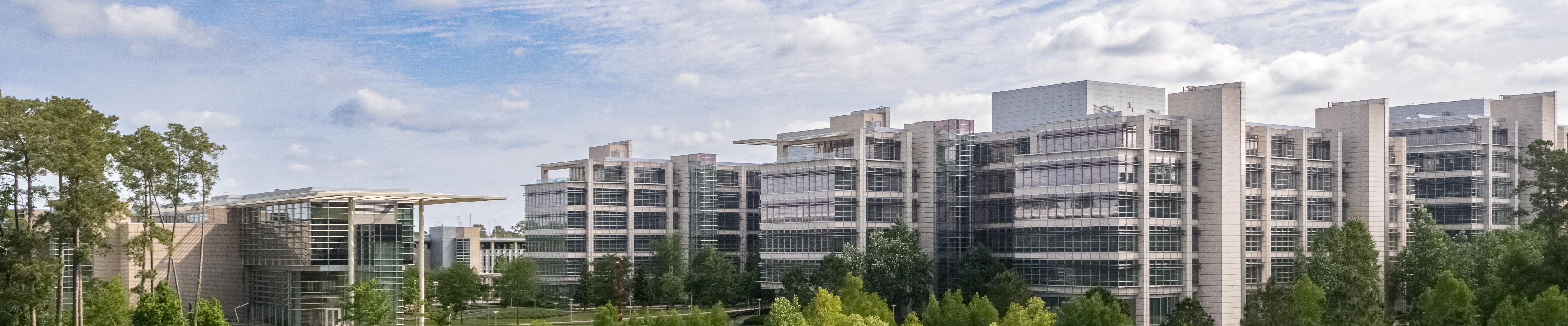 A modern office complex with several large, glass-fronted buildings situated behind a scenic pond. The architecture features clean lines and extensive use of windows, allowing natural light to flood the interiors. The foreground is dominated by the pond, surrounded by lush greenery and trees, providing a tranquil, park-like setting. The sky is partly cloudy, adding to the serene atmosphere. There are walking paths and outdoor spaces visible, suggesting the area is designed to encourage outdoor activity and relaxation.