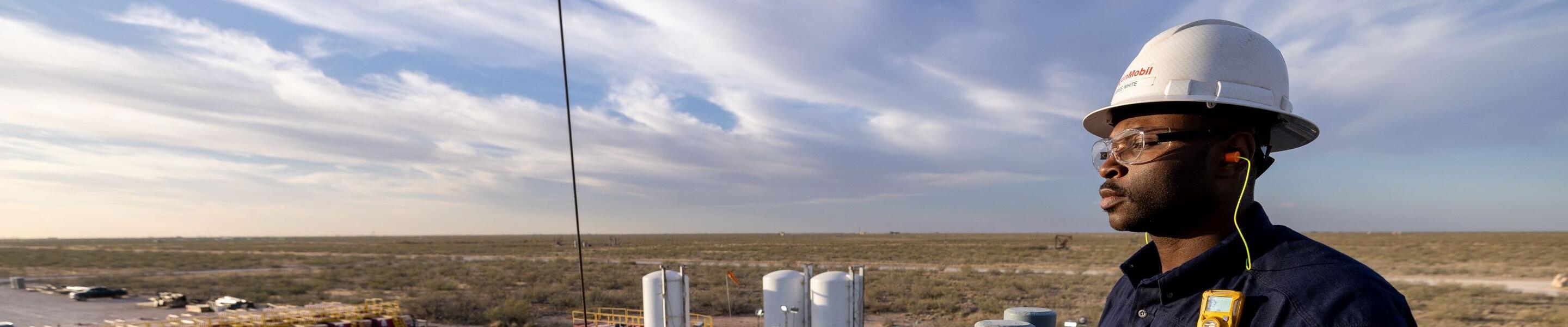 An industrial scene at what appears to be an oil or gas site. A worker wearing a hard hat, protective gloves, and safety gear is standing on a platform, overlooking the site. The person is wearing a shirt with the 