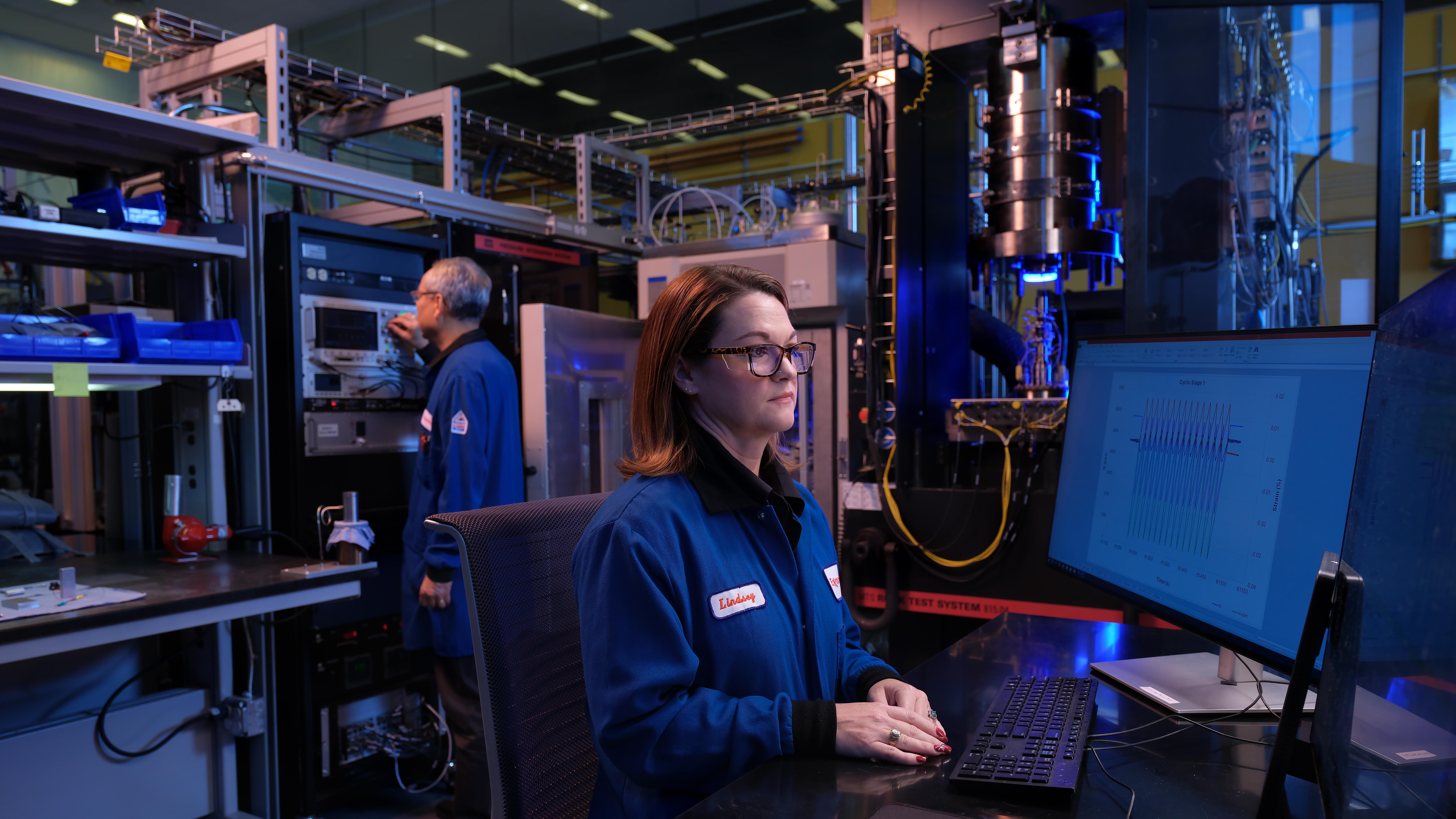 A high-tech lab or manufacturing facility with two people wearing blue lab coats working among racks of equipment and cabling.
