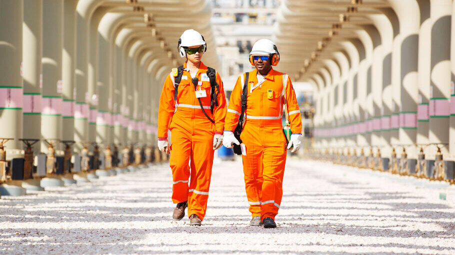 Two workers dressed in bright orange protective coveralls walking side by side in an industrial facility. They are wearing white helmets, gloves, and ear protection, and each carries a backpack. The scene is set between large parallel pipes, which appear to run the length of the facility, suggesting it may be related to oil, gas, or chemical processing.