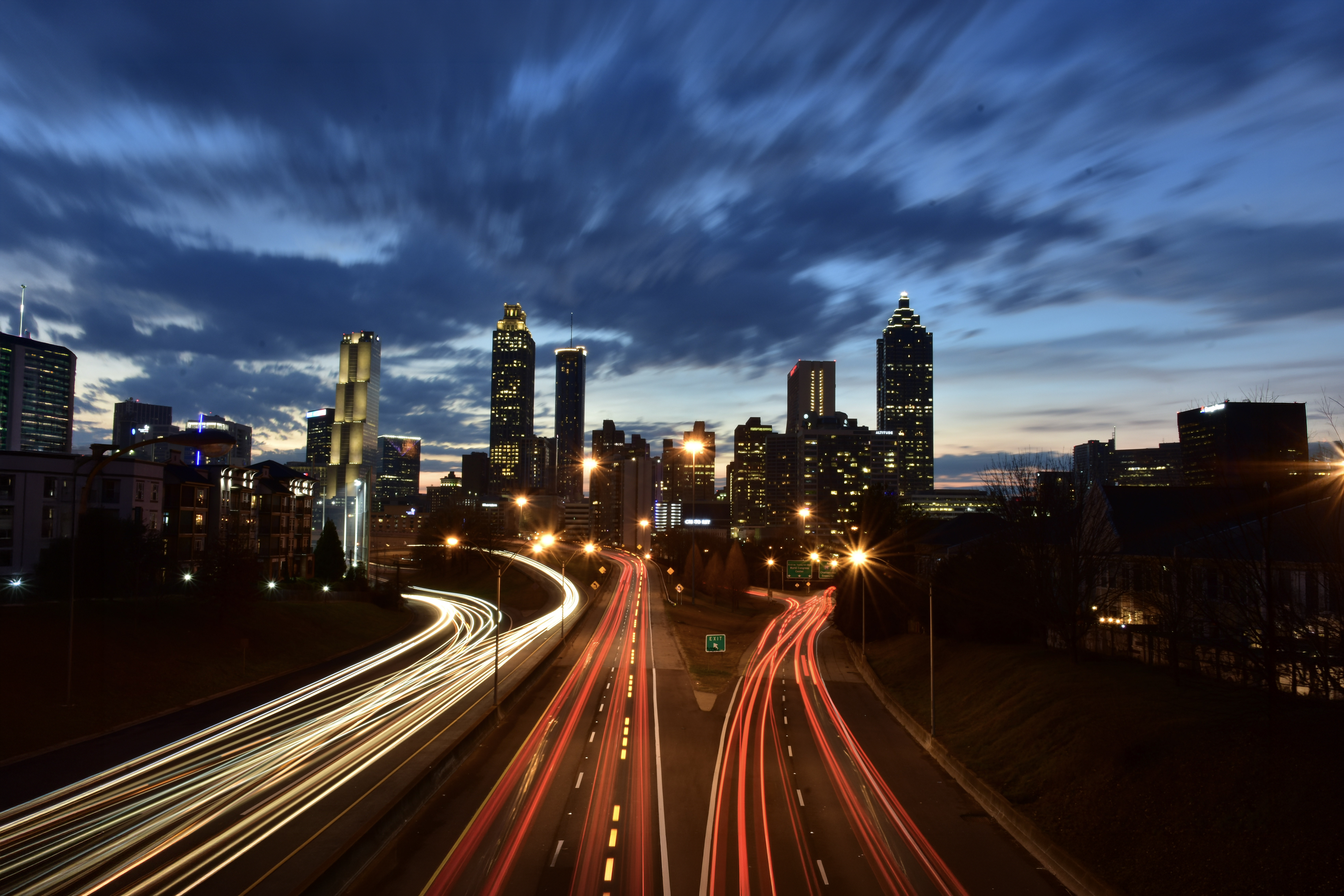 A dusk cityscape shows a modern skyline with tall, illuminated buildings under streaky blue clouds. In the foreground, a multi-lane highway curves into the city, captured with long exposure so headlights form white streams and taillights form red ribbons. Streetlights glow amber along the road, and the horizon still holds a faint band of sunset light. The scene feels dynamic and energetic as traffic flows toward the downtown core.