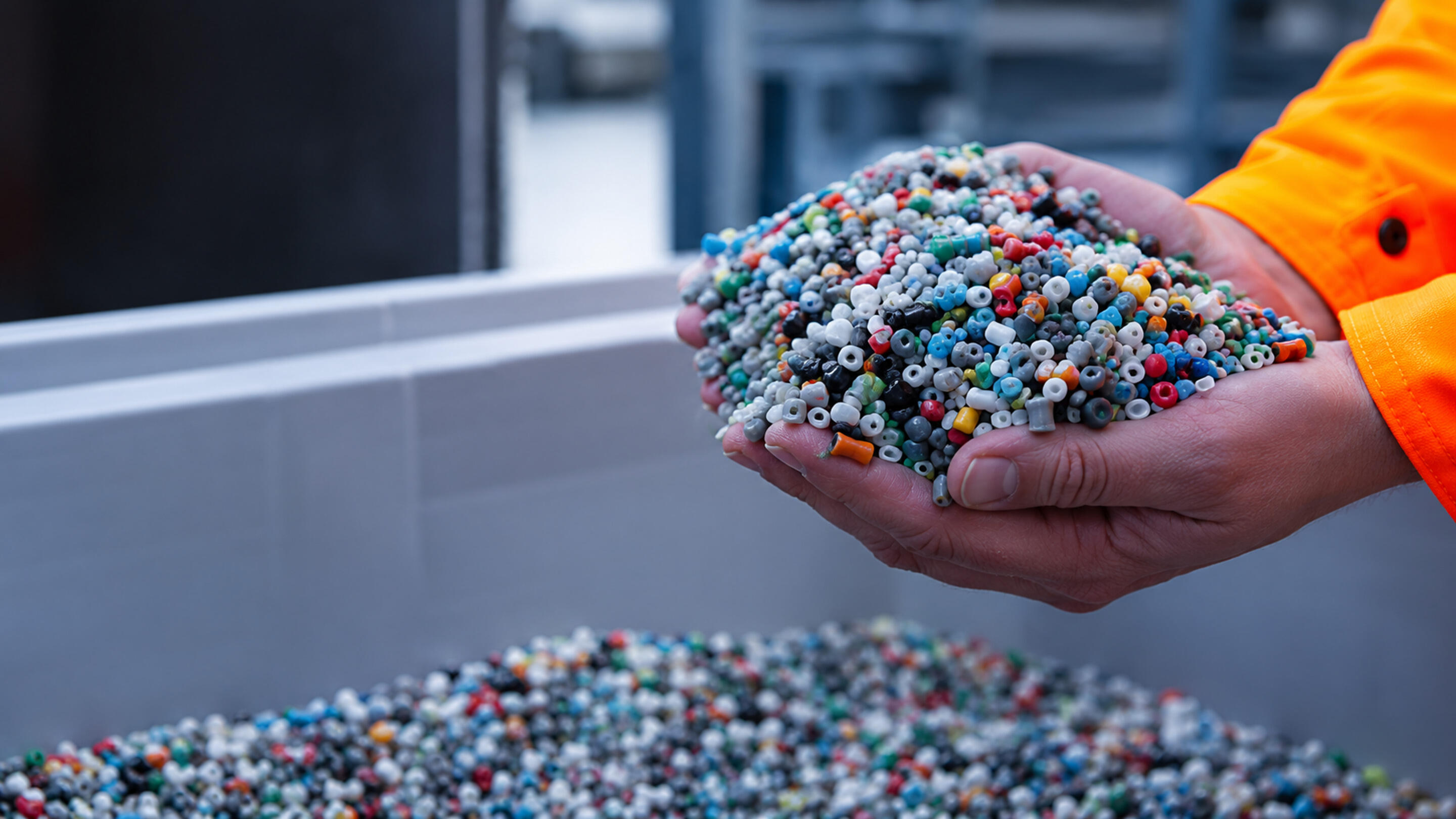 A person wearing an orange sleeve holds a large handful of small, multicolored plastic pellets or beads.