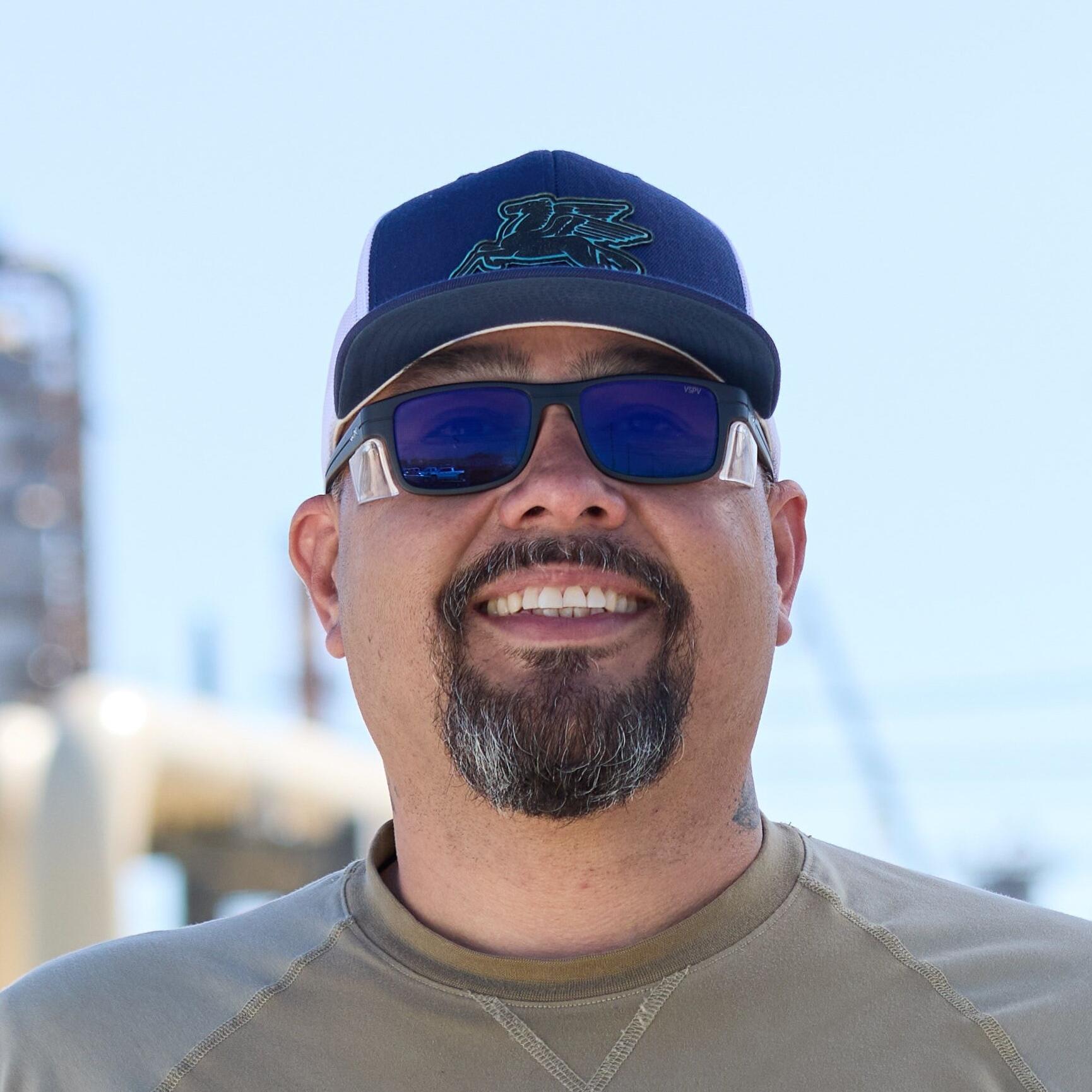 A man wearing sunglasses, a cap, and a tan long-sleeve shirt stands smiling in an industrial area. He holds a white hard hat with the ExxonMobil logo by his side, and his shirt also has red ExxonMobil text.