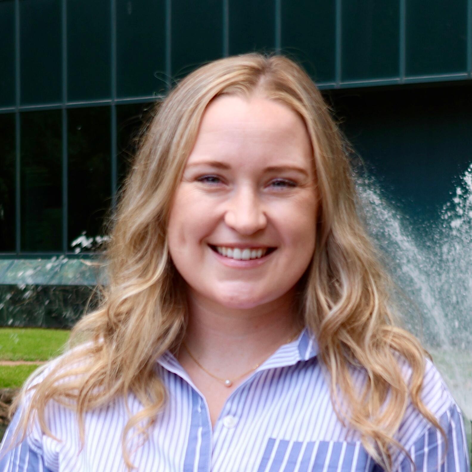 A smiling person stands outdoors in front of a modern glass office building and a small pond with a fountain.