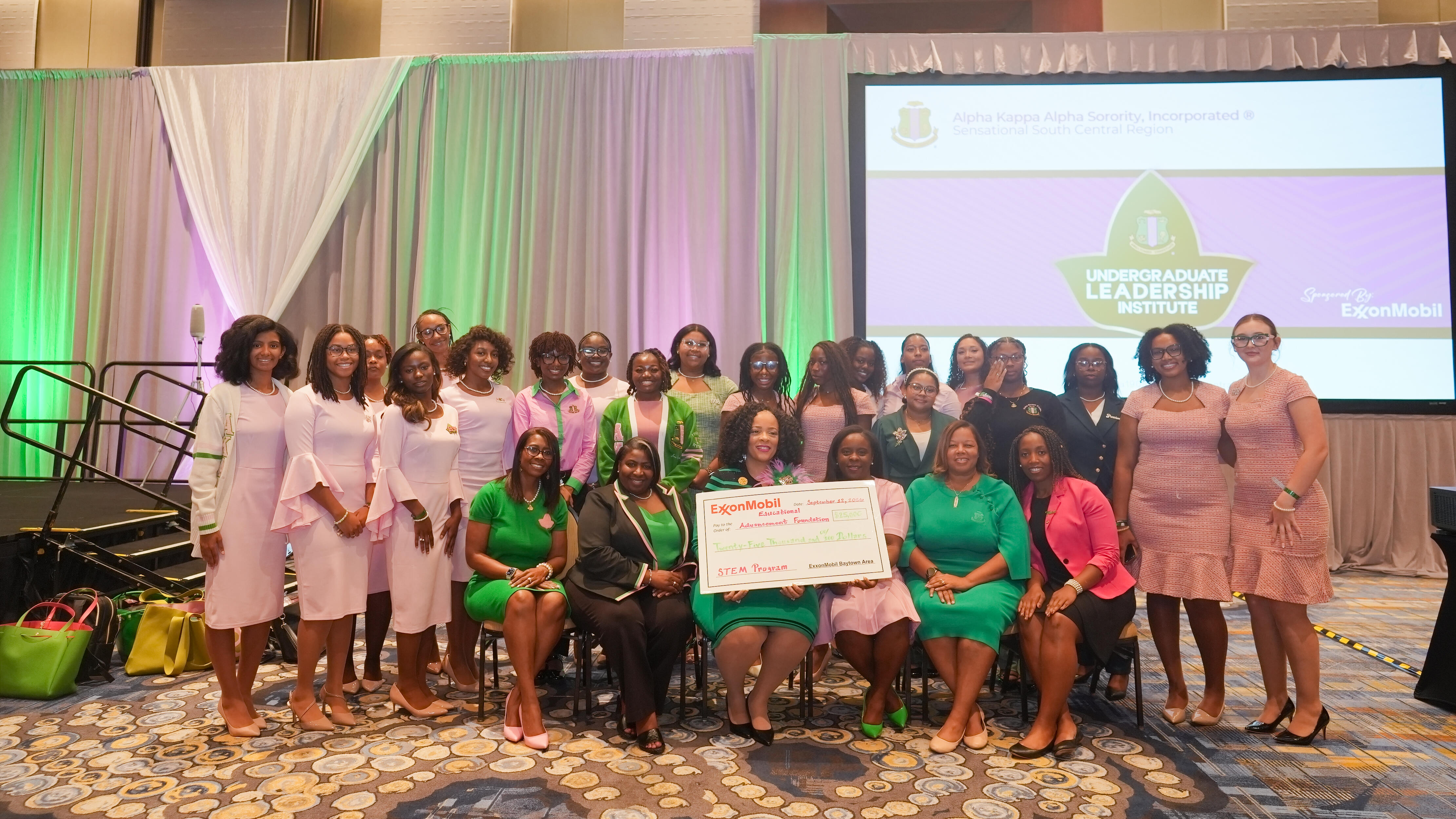 A large group of women pose together in a ballroom, many wearing coordinated pink and green outfits.