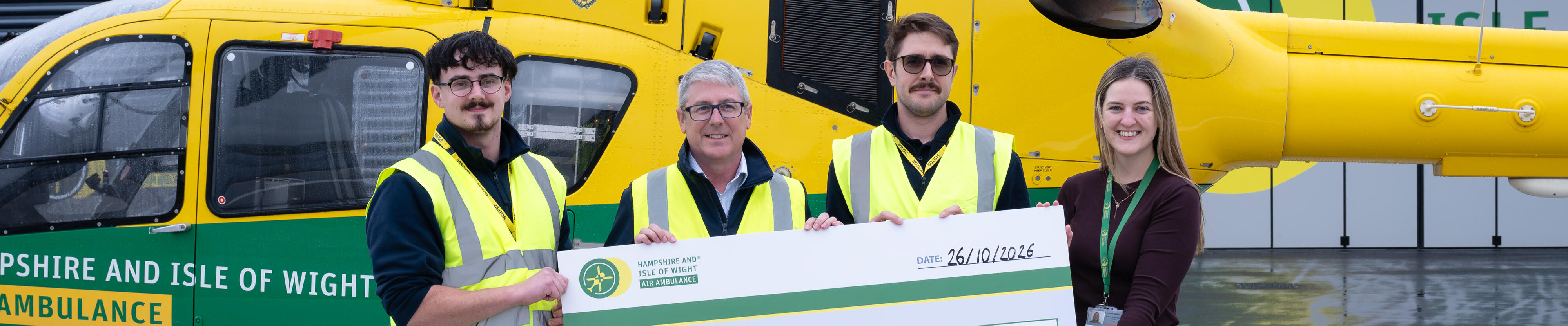 Four people stand in front of a Hampshire and Isle of Wight Air Ambulance helicopter, holding a ceremonial £20,000 donation check from ExxonMobil.