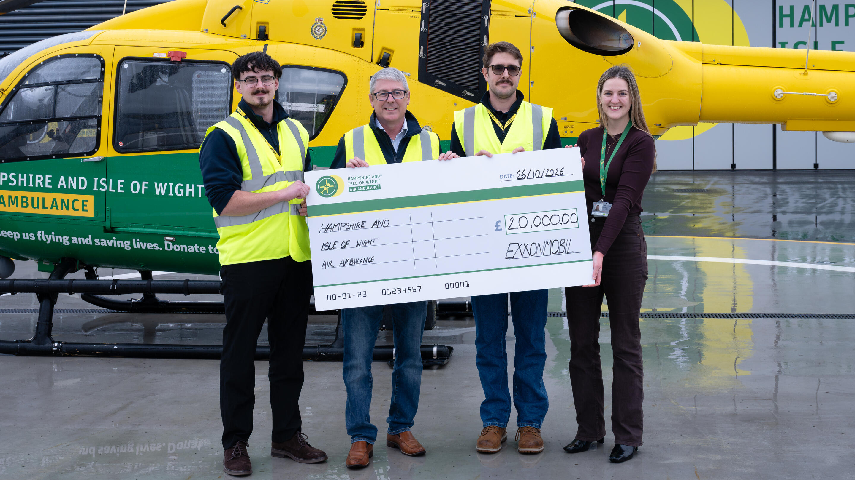 Four people stand in front of a Hampshire and Isle of Wight Air Ambulance helicopter, holding a ceremonial £20,000 donation check from ExxonMobil.