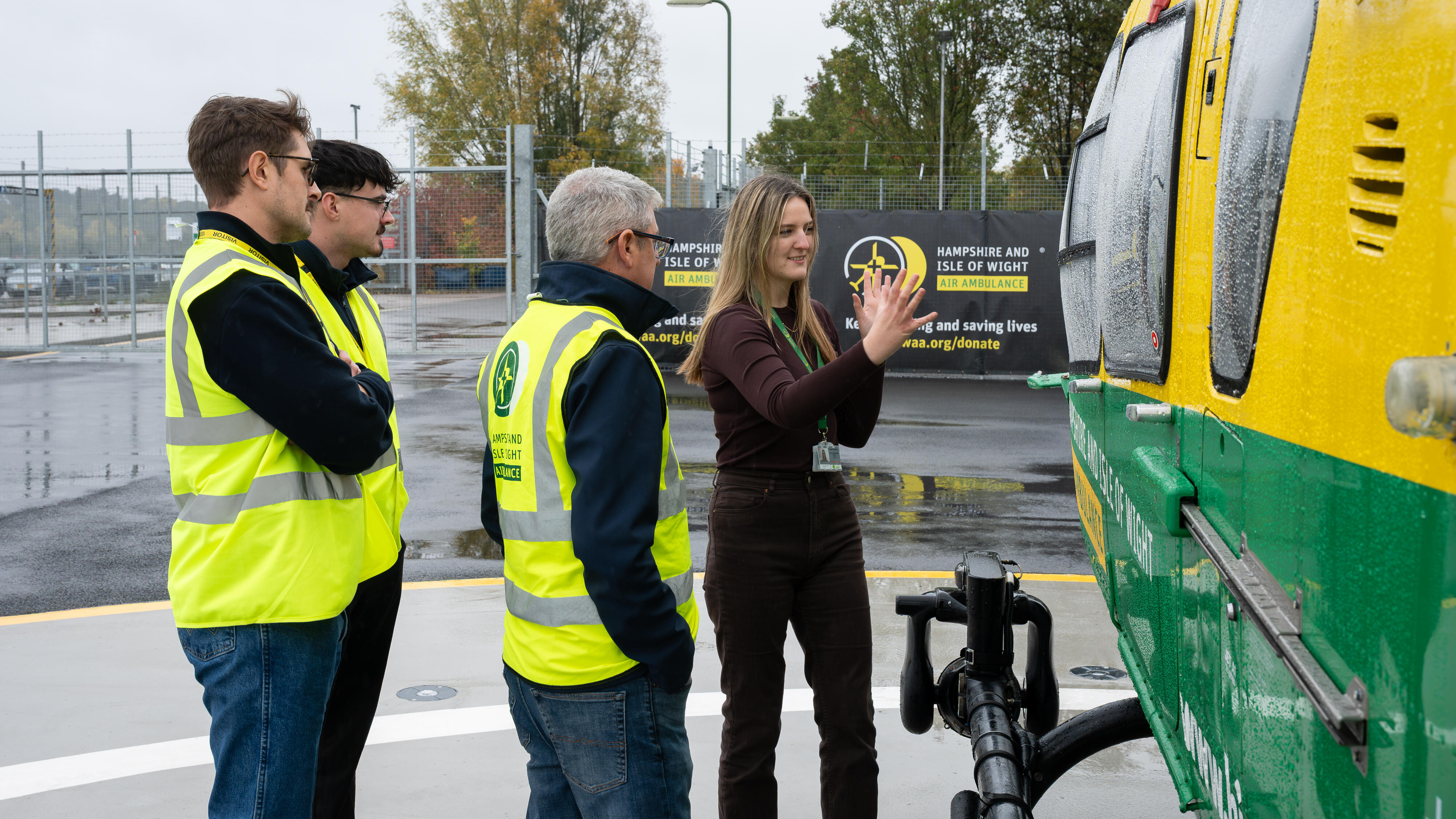 Four people stand beside a Hampshire and Isle of Wight Air Ambulance helicopter on a wet helipad, inspecting the aircraft during a site visit.