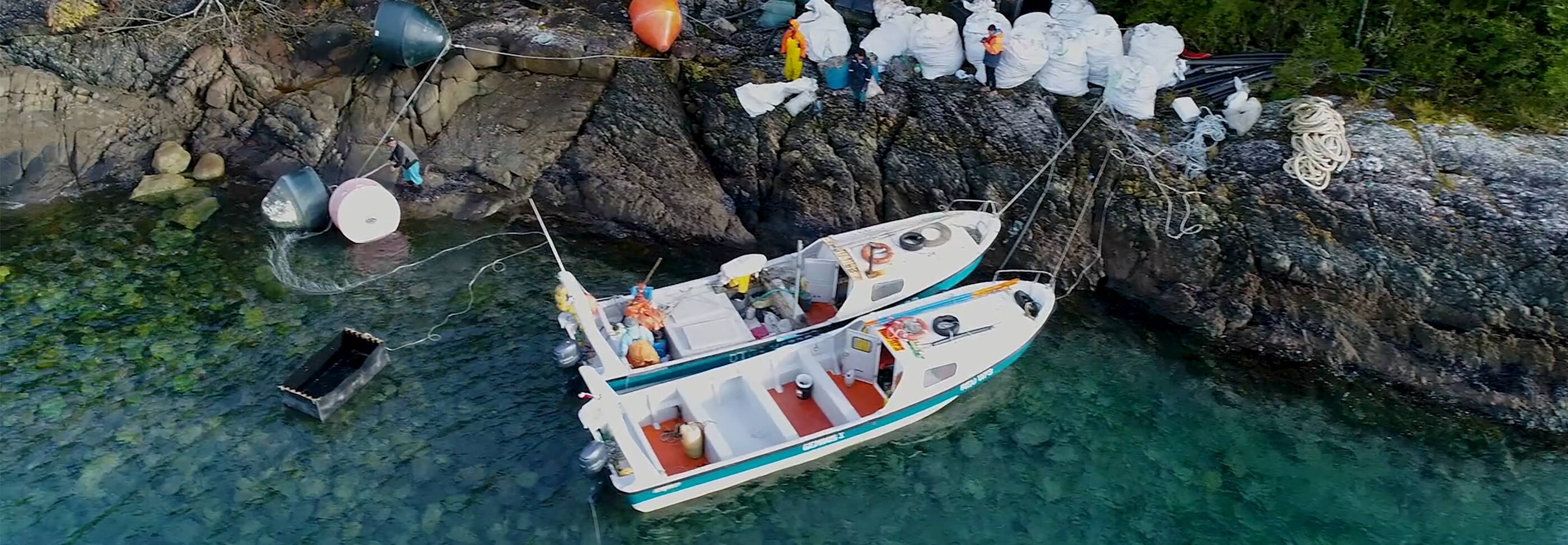 Two boats in the water tied to the shore, with bags full of recycles lining the shoreline.