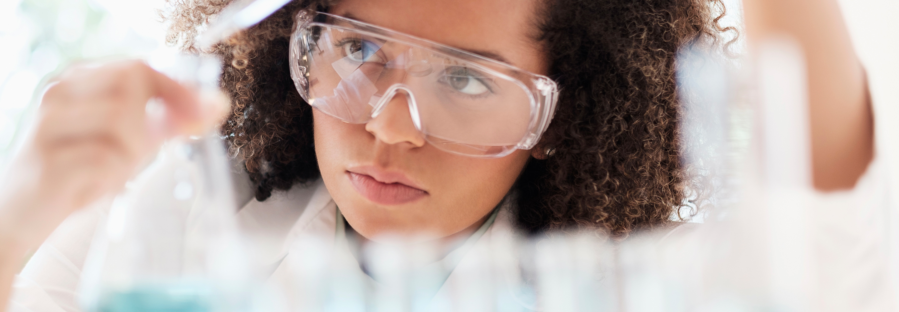 A female scientist with goggles in her lab examining substances in beakers.