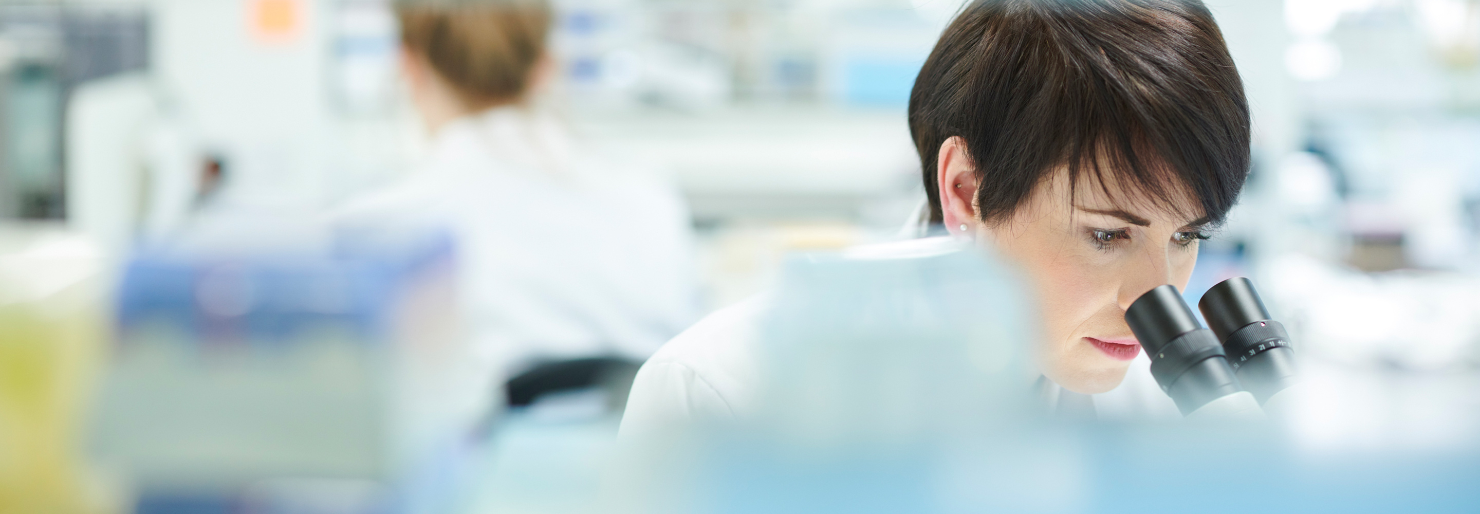 Female scientist in her lab looking into a microscope with another scientist in the background.